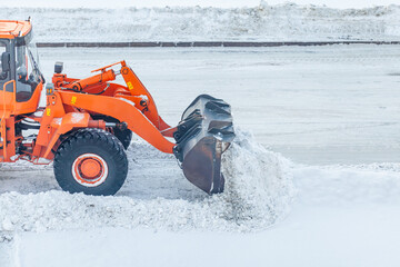 Fototapeta na wymiar Big orange tractor cleans up snow from the road and loads it into the truck. Cleaning and cleaning of roads in the city from snow in winter
