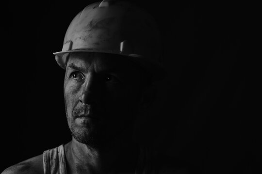 Dirty face of coal miner on a black background. Head of tired mine worker in a hard hat. Black and white photographic portrait.