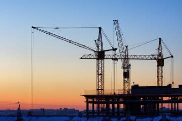 Construction cranes are building a house against the background of sunset and dark sky