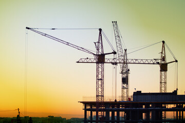 Construction cranes are building a house against the background of sunset and dark sky