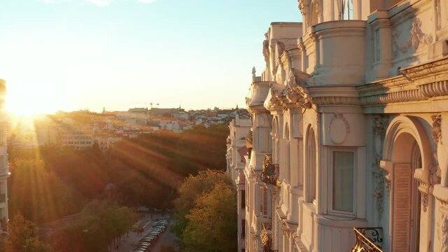Forwards fly along historic palace with decorated facade. Elevated view of buildings in city. Illuminated by bright setting sun.