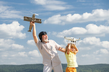 Young grandson and old grandfather with plane and quadcopter drone over blue sky and clouds background. Men generation granddad and grandchild. Elderly old relative wit child.