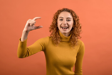 Young and attractive redhead Caucasian girl in orange jumper showing small size with her fingers...
