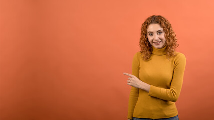 Young and attractive redhead Caucasian girl in orange jumper pointing her finger at copy space for advertising isolated on orange studio background.