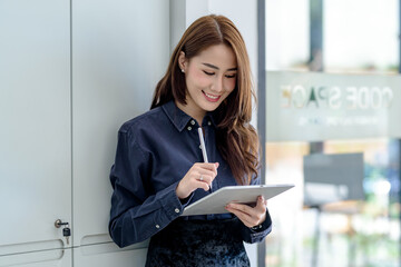 Front view happy successful young Asian woman holding a pen using a tablet standing at the office.