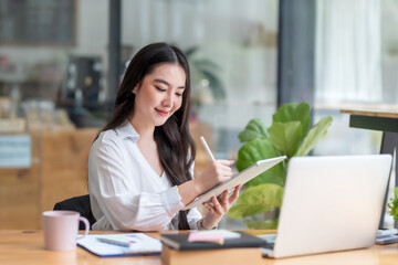 Fototapeta premium Portrait of smiling beautiful Asian businesswoman sitting enjoy the idea with tablet and laptop at office..