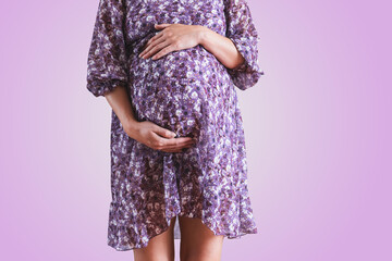 Pregnant woman in dress holds hands on belly on a violet background.