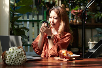 Young woman sitting at table in outdoor cafe, looking at compact mirror and applying liquid lipstick