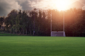 Tall goal post for Irish National sports on a training pitch against dark trees at sunset. Sun flare. Copy space. Camogie, hurling, rugby and Gaelic football playground. Calm mood.