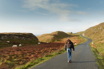 Young teenager girl walking uphill on a small asphalt twisted road. Sheep grazing grass on a field off the road. Travel and reaching goals concept. Tourist exploring nature scene