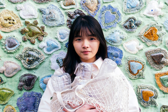 Young woman holding mesh bag with plastic bottles in front of carved wall