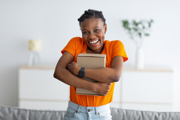 Portrait of smiling black female buyer hugging cardboard box, receiving parcel, satisfied with great purchase at home
