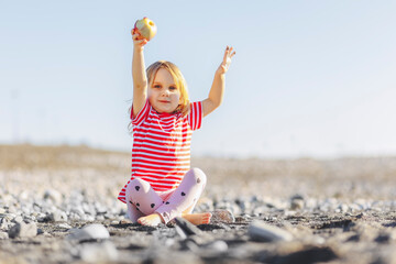 Girl with arms raised holding apple sitting at beach on sunny day