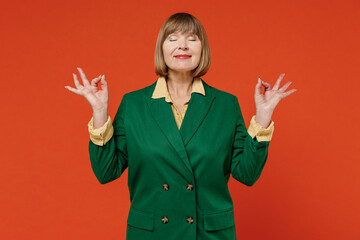 Elderly spiritual tranquil woman 50s wearing green classic suit hold spreading hands in yoga om aum gesture relax meditate try to calm downisolated on plain orange color background studio portrait
