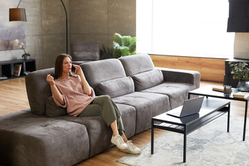 Obraz premium Young woman sitting on sofa and talking on her mobile phone in modern living room at home