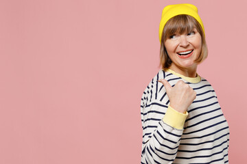 Elderly stylish woman 50s in striped shirt yellow hat point thumb finger aside on workspace area mock up area isolated on plain pastel light pink background studio portrait. People lifestyle concept.