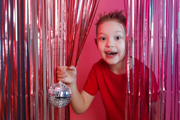 Little cute boy with shows a disco ball and smile on the background of a foil curtain.