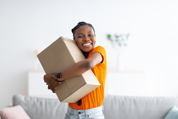 Joyful black woman embracing carton parcel, receiving desired delivery, getting online order at home