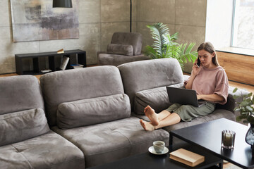 Obraz premium Young freelancer lying on the sofa, she working at home typing on laptop and having a conversation on mobile phone