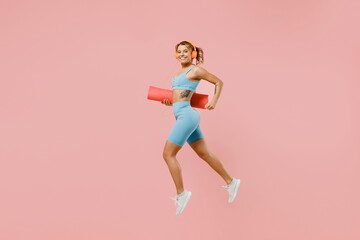 Fototapeta na wymiar Full body young strong sporty fitness trainer instructor woman wear blue tracksuit headphones spend time in home gym hold mat jump high isolated on pastel plain pink background. Workout sport concept.