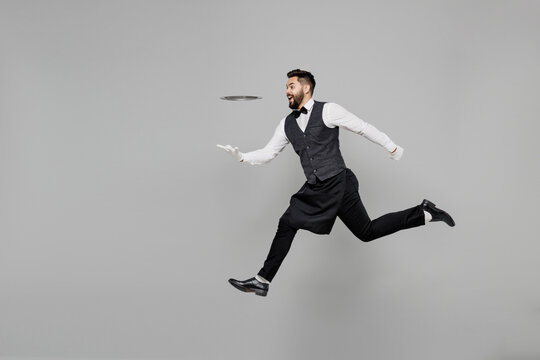 Full Body Young Male Waiter Butler Man 20s Wear White Shirt Vest Elegant Uniform Work At Cafe Jump High Hurrying Up Run Hold Metal Tray Isolated On Plain Grey Background . Restaurant Employee Concept.