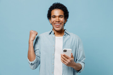 Young black curly man 20s years old wears white shirt hold in hand use mobile cell phone doing winner gesture celebrate clenching fists isolated on plain pastel light blue background studio portrait.