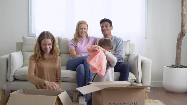 Happy volunteer caucasian family unpack, packing stuff clothes in cardboard box donate to needy people while sitting in living room at home in winter day. Donation and charity concept.