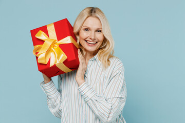 Elderly woman 50s wearing casual striped shirt looking camera hold red present box with gift ribbon...