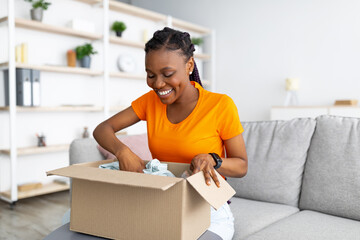 Online delivery service. Cheerful African American woman unpacking parcel box with purchase at home