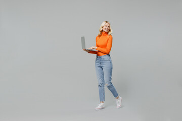 Full body elderly smiling happy blonde woman 50s in orange turtleneck hold use work on closed laptop pc computer look aside on workspace area mock up isolated on plain grey color background studio