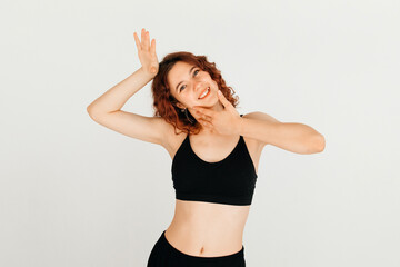 Fototapeta na wymiar Cheerful woman with curly red hair smiling, posing with her hand near her face in black sport top, standing at white background