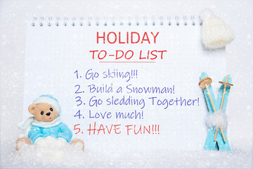 Notepad with to-do list: skiing, making a snowman, sledding, loving, having fun and a toy Teddy bear, blue skis, on snow