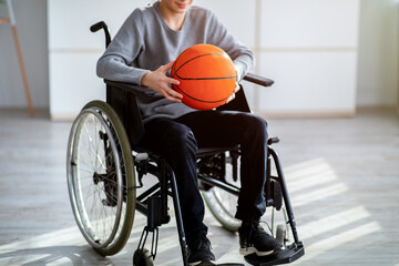 Cropped view of disabled teenager in wheelchair holding basketball at home. Impairment and sports...