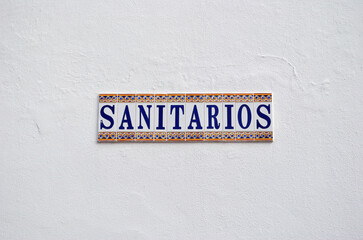 Close Up of Decorative Ceramic Tile Sign 'Sanitarios' on White Wall Translates as 'Toilets'