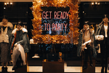 Neon shiny holiday signboard on boutique showcase with text Get ready for party and female mannequins