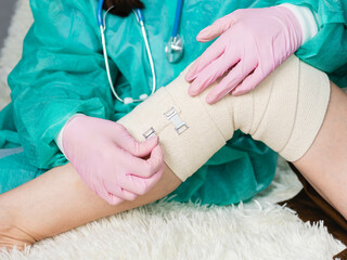 A doctor wearing medical gloves wraps an elastic bandage around the patient's leg and knee. The...