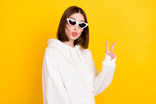 Photo of romantic lady show v-sign send air kiss wear sunglass white hoodie isolated yellow color background