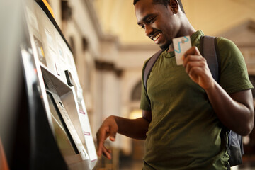 Smiling African man using ATM machine. Happy young man withdrawing money from credit card at ATM. - obrazy, fototapety, plakaty