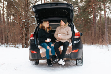 Happy young couple in love having rest in trunk of car in winter snowy forest, outdoors. Man and...