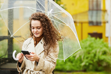 Horizontal medium portrait of young woman wearing trench coat standing outdoors under umbrella on...