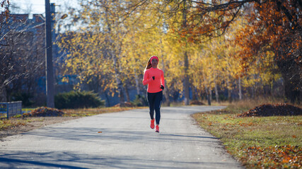 Young fitness woman running on the road in the cold morning in winter. Sporty girl in orange long-sleeve jogging in the sunny morning