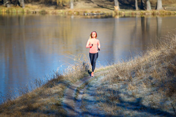 Fitness girl jogging in park near pond in the frosty morning. Running woman training near the lake in cold weather
