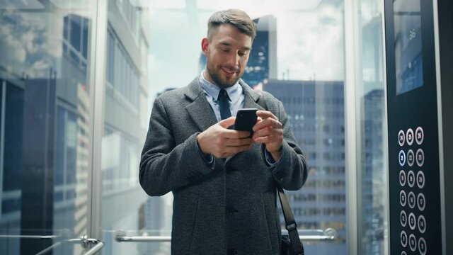 Successful Businessman Riding Glass Elevator to Office in Modern Business Center. Handsome Happy Man Smile while Using Smartphone, Write Text Message, Check Social Media and Work Emails in a Lift.