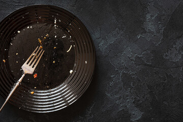 Dirty glossy black plate and a fork on a black textured background with copy space. Dirty and empty dishes after meals and holidays. A plate after a delicious pasta. Flat lay