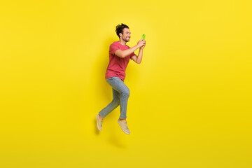 Full size photo of nice beard millennial guy jump look telephone wear red t-shirt jeans sneakers isolated on yellow background