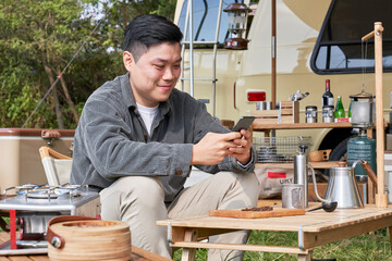 A young man healing himself by operating a smartphone in front of a camping car at the campsite