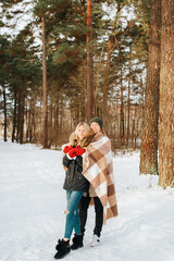 Fototapeta premium Happy couple in love wrapped in blanket hugging in winter forest on sunny snowy day. Caucasian young men and women enjoying walk together in nature. Valentine's day, family weekend, romantic lifestyle