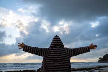 White man at the edge of the sea wearing a hooded coat and arms outstretched in hugging pose.