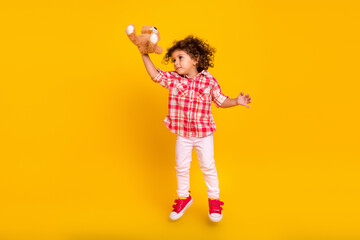Photo of pretty sweet school girl dressed checkered clothes jumping holding toy looking empty space isolated yellow color background