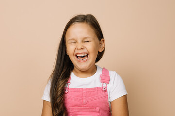 Portrait of child with fully opened mouth, closed eyes and excited smile with teeth looking at camera wearing bright pink jumpsuit and white t-shirt on beige background.